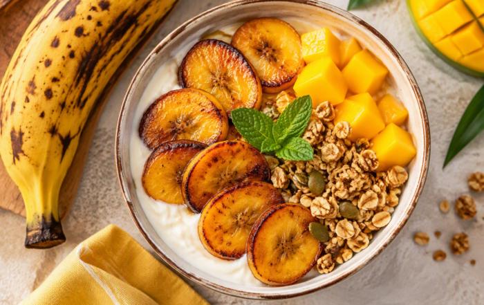 Bol tropical vu de dessus composé de yaourt grec, de rondelles de banane plantain caramélisées, de dés de mangue, de granola et de feuilles de menthe, accompagné d’une banane plantain mûre.