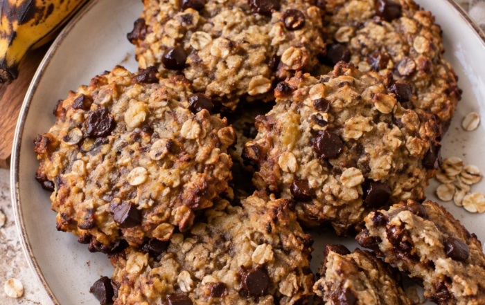 Vue de dessus de cookies maison à la banane plantain très mûre, aux flocons d’avoine et aux pépites de chocolat noir, servis dans une assiette avec des bananes plantains en arrière-plan.
